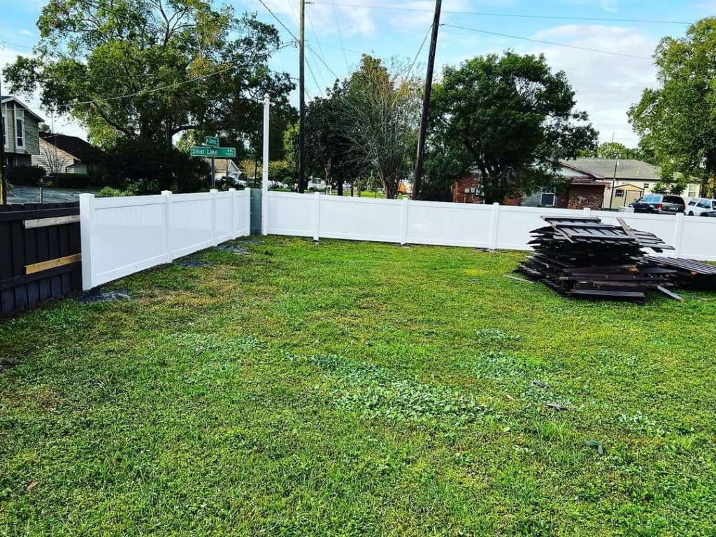 Backyard fresh white vinyl privacy fence corner removed dark wood planks stacked grass street sign leafy neighborhood sky.