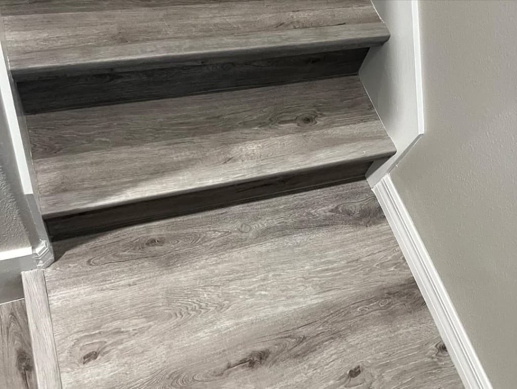 Stair tread grey vinyl plank nosing trim white PVC baseboards corner transition beige wall bright lighting detail.