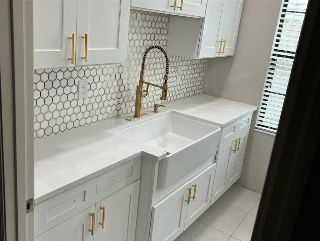 Farmhouse sink gold faucet white hex backsplash with gold grout lines shaker drawers window blinds countertop quartz.