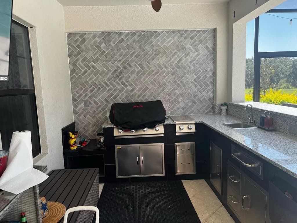 Outdoor grill station with recessed grey herringbone tile, granite countertop, grill cover, and backyard view beyond.