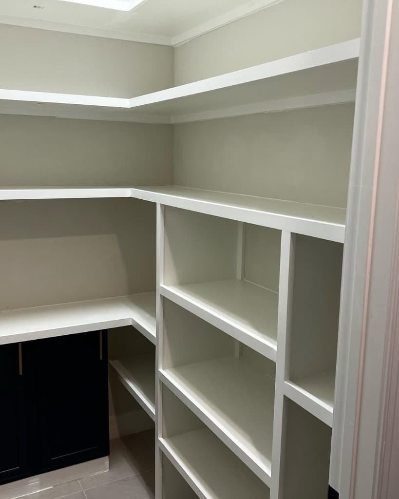 Walk-in pantry corner with painted white shelves, cubby divider wall, navy base cabinet doors, and gold pulls.